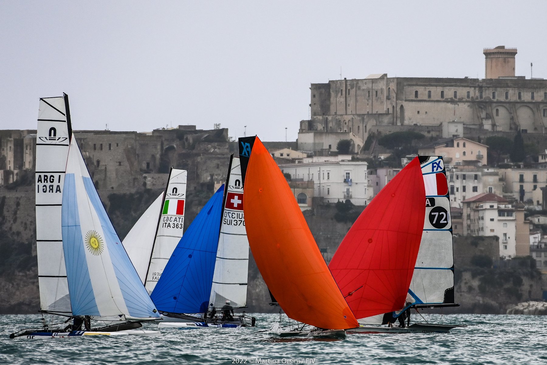 Barche a vela con spinnaker colorati davanti al castello e alle case di un borgo costiero