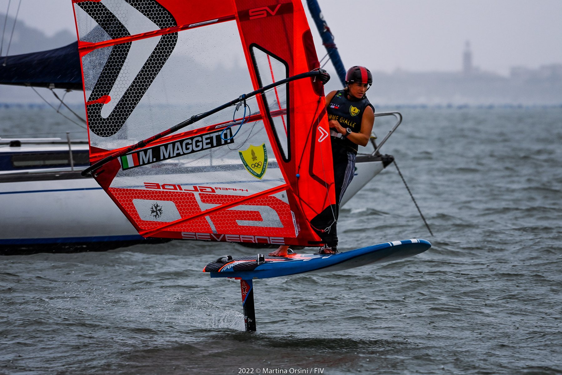 Windsurfista in volo su foil con vela arancione sotto la pioggia