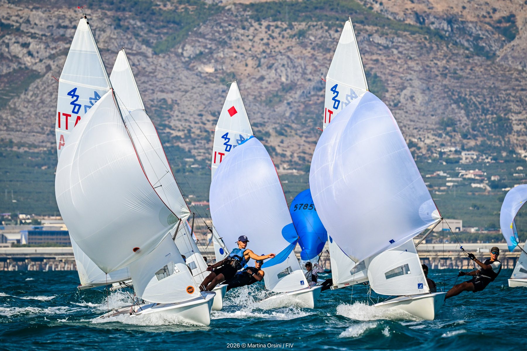 Imbarcazioni a vela bianche in regata davanti a una costa montuosa