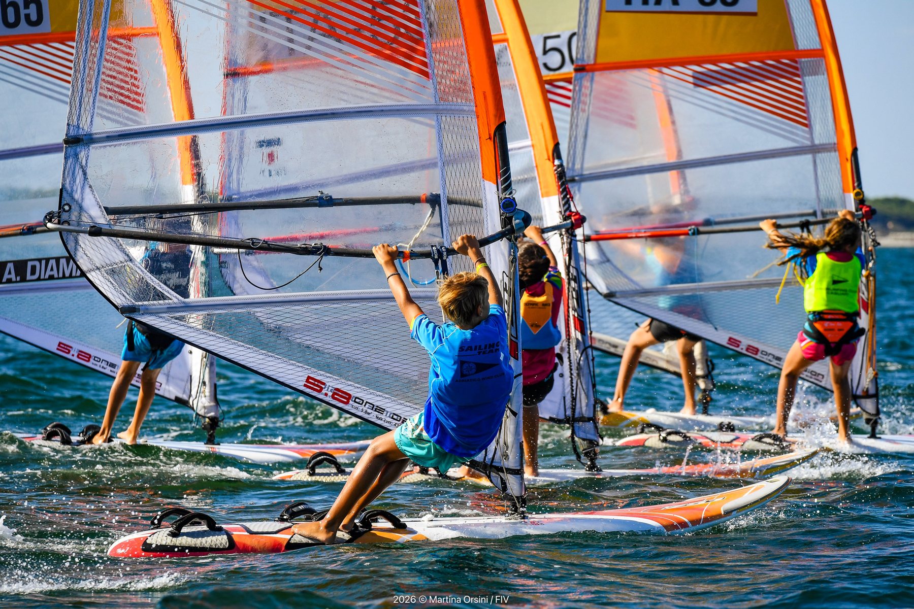 Gruppo di giovani windsurfisti con vele arancioni durante una regata